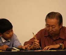 周老菩薩的孫子在一旁看著爺爺用毛筆寫出一句句的靜思語。【攝影者：何义雄】