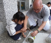 志工王志勤握著白薈恩小朋友手中的鐵鎚，在鐵罐上敲打出洞口。【攝影者：張秋虹】
